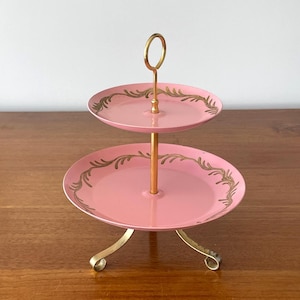 May include: A two-tiered pink serving tray with gold-colored accents. The trays have a gold floral design around the edges and a gold handle. The tray is supported by three curved gold legs.