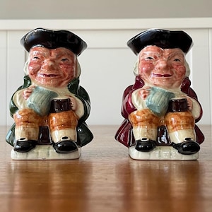 May include: Two ceramic Toby jugs, each depicting a smiling man in a tricorn hat and holding a mug. One wears a green coat, the other a burgundy coat. The jugs have brown, tan, and black accents.