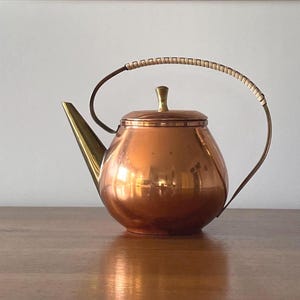 Puede incluir: Una tetera de cobre con boquilla y asa de latón. El asa está envuelta con un material de color claro. La tetera tiene un cuerpo redondo y una tapa con un pomo de latón. La tetera está sobre una superficie de madera.