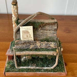 May include: A miniature rustic bench with a stone chimney and a small house. The bench is made of wood and bark, with a green mossy base. A small card reads "The Country Charm Collection".