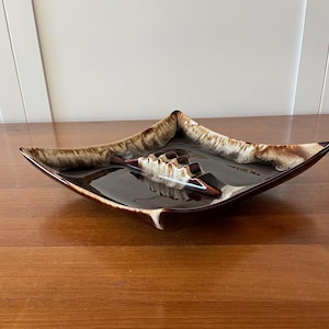 May include: A square ashtray with a dark brown glossy finish and a lighter brown and cream-coloured rim. The ashtray has a built-in cigarette rest and is made of ceramic or a similar material. The ashtray is placed on a wooden surface.