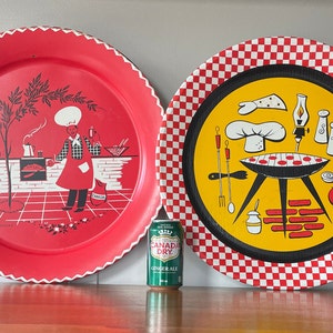 Op de afbeelding: Twee vintage metalen serveerschalen met rood-wit geblokte randen. Een schaal heeft een rode achtergrond met een zwart-witte illustratie van een kok die in een keuken kookt. De andere schaal heeft een gele achtergrond met een zwart-witte illustratie van een grill met hotdogs, een koksmuts en andere grillbenodigdheden.
