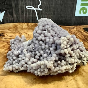 Peut inclure: Un amas de calcédoine botryoïdale violet clair, ressemblant à des raisins, est posé sur une surface en bois. La pierre a une texture bulleuse et des nuances de lavande. Le fond comprend un panneau en bois et une partie d'une enseigne.