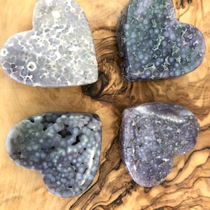 Peut inclure: Quatre pierres en forme de cœur avec une surface texturée et bulleuse. Les pierres sont de différentes nuances de violet et de gris, avec quelques accents verts. Elles sont disposées sur une surface en bois brun clair.