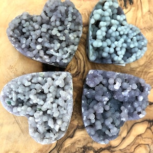 Peut inclure: Quatre géodes d'améthyste en forme de cœur avec une texture botryoïdale et bulleuse. Les géodes varient en nuances de violet et de gris, sur une surface en bois brun clair. Les formes de cœur sont clairement définies, mettant en valeur les formations naturelles des cristaux.