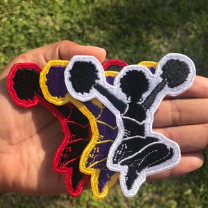 May include: Three embroidered patches featuring a cheerleader silhouette in a jumping pose with pom-poms. The patches are layered, showcasing black figures with white, yellow, and red stitched borders, each with a distinct colored edge.