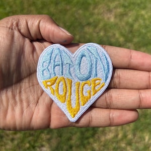 May include: A heart-shaped embroidered patch featuring the text Baton Rouge. The top portion of the heart displays the word Baton in light blue thread, while the bottom portion features the word Rouge in bright yellow thread on a white background.