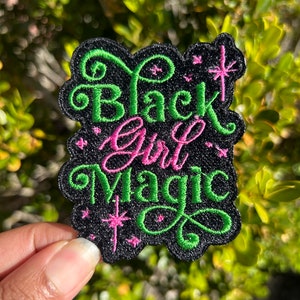 May include: Black Girl Magic embroidered patch with green lettering on a black background. The patch features a pink star and a pink swirl in the lettering.