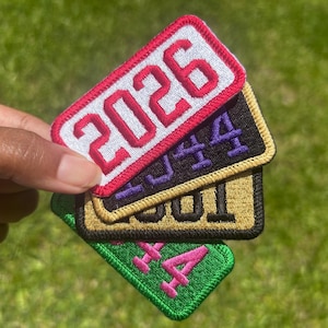 May include: A stack of four rectangular embroidered patches with stitched borders. The top patch is white with red 2026 text, followed by a black patch with purple text, a gold patch with black text, and a green patch with pink text.