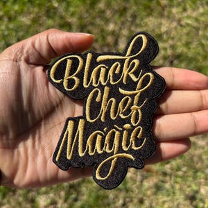 May include: Black embroidered patch with the words "Black Chef Magic" in gold script lettering. The patch is black with gold stitching and is held in a hand against a blurred green background.