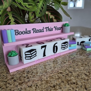 May include: A pink wooden desk calendar with the text "Books Read This Year" and "Randomly Me". The calendar displays the numbers 4, 7, and 6, along with book icons. It includes miniature book and succulent decorations.