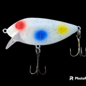 Puede incluir: Un señuelo de pesca blanco con manchas rojas, azules y amarillas. El señuelo tiene un labio de plástico transparente y dos anzuelos triples. El señuelo está diseñado para atraer peces.