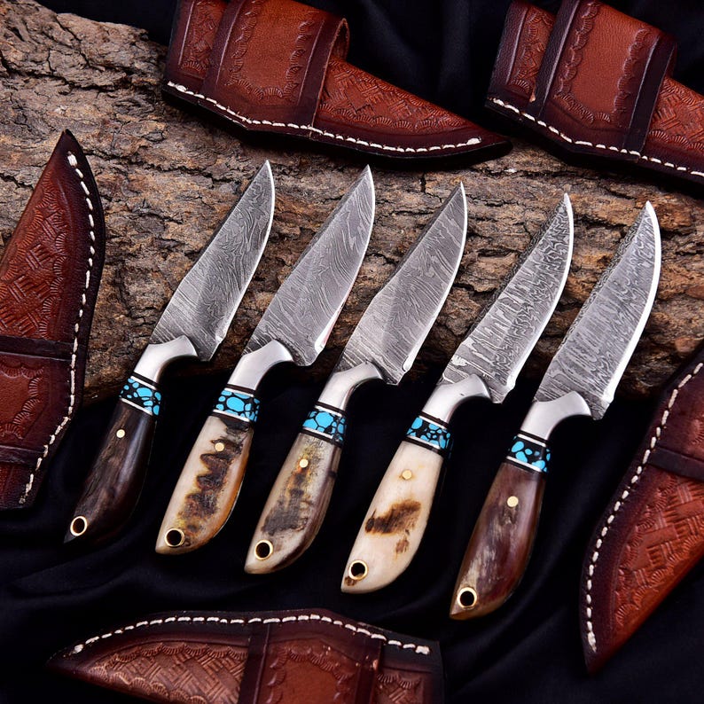 May include: A collection of five Damascus steel knives with decorative handles. The handles feature a combination of dark wood, bone, and turquoise accents. Each knife is paired with a brown leather sheath, showcasing a detailed embossed pattern. The knives are arranged on a dark surface.