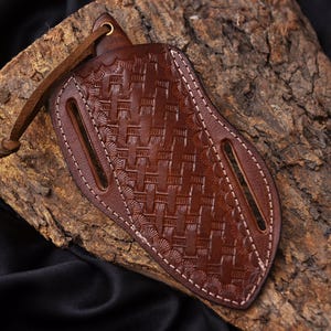 May include: A brown leather knife sheath with a woven pattern and white stitching. The sheath has two slots and a leather strap with a brass grommet. The leather is a rich, dark brown color.