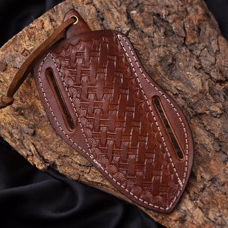May include: A brown leather knife sheath with a woven pattern and white stitching. The sheath has two slots and a leather strap with a brass grommet. The leather is a rich, dark brown color.