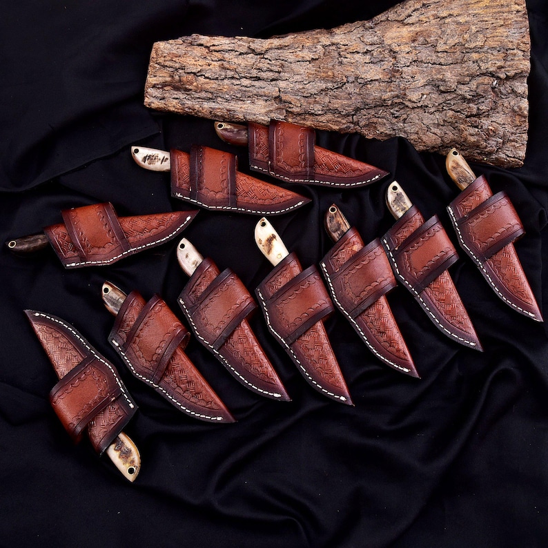 May include: A collection of handcrafted knives with brown leather sheaths. The knives have bone handles and are displayed on a black surface with a piece of wood in the background. The sheaths are intricately stitched.