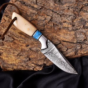 May include: A handcrafted knife with a patterned blade and a bone handle. The handle features a blue and silver accent. A brown leather cord is attached to the handle. The knife is displayed on a piece of wood.