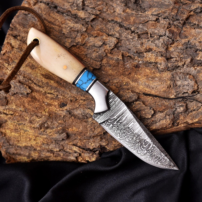 May include: A handcrafted knife with a patterned blade and a bone handle. The handle features a blue and silver accent. A brown leather cord is attached to the handle. The knife is displayed on a piece of wood.