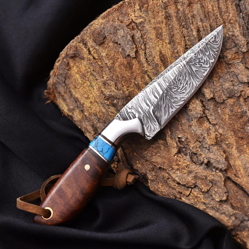 May include: A handcrafted knife with a patterned steel blade and a wooden handle. The handle features a turquoise accent and a leather lanyard. The knife is displayed on a wooden surface against a black background.