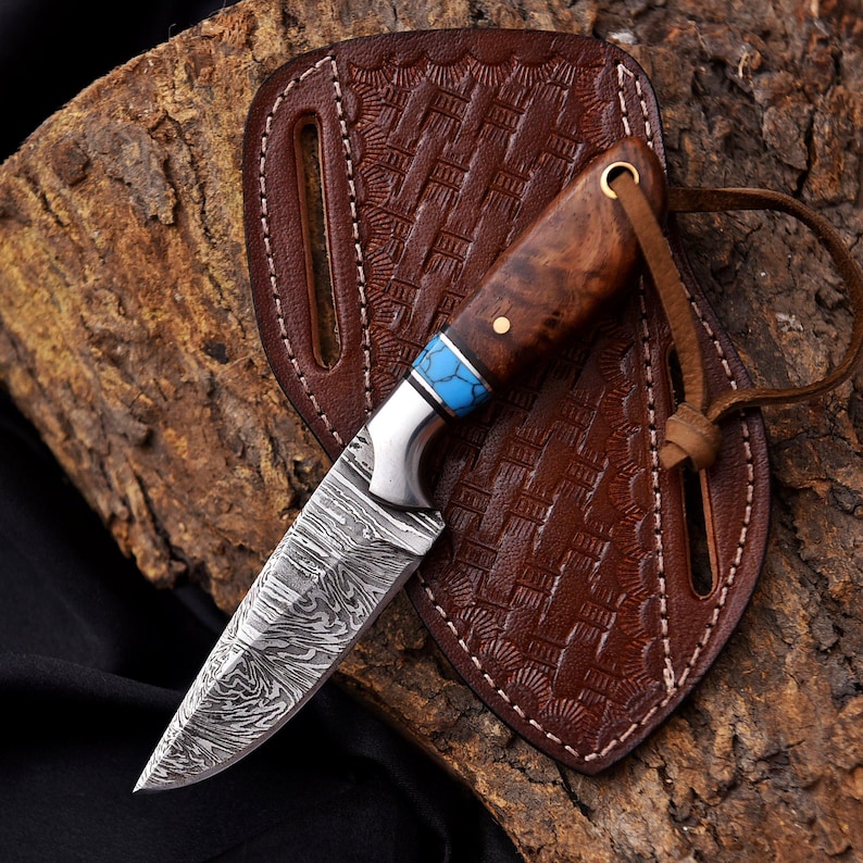 May include: A handcrafted knife with a patterned steel blade and a wooden handle with blue accents. The knife rests on a brown leather sheath with a textured design, and a leather strap. The knife is displayed on a piece of wood.