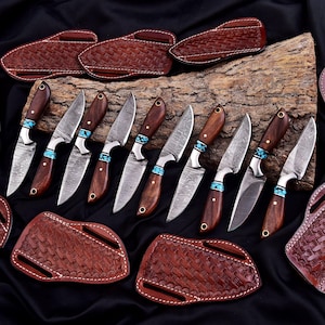 May include: An assortment of handcrafted knives with wooden handles and turquoise accents. The blades have a unique pattern, and each knife comes with a brown leather sheath. The knives are arranged on a dark surface.