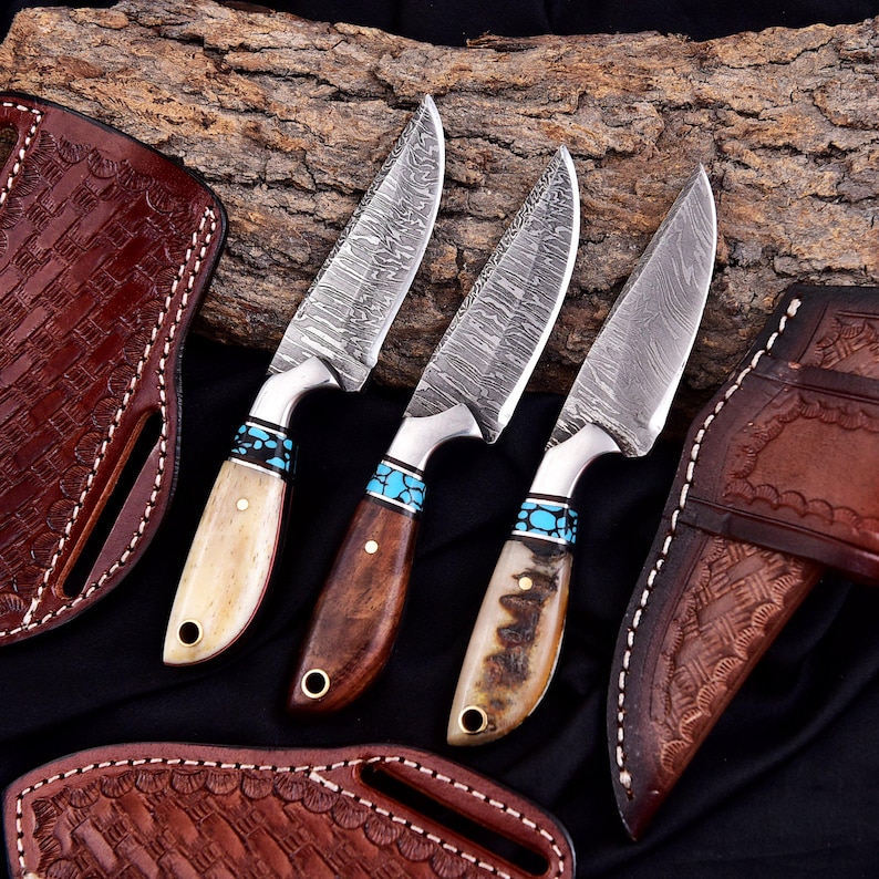 May include: Three Damascus steel knives with turquoise accents and wooden or bone handles. The blades have a unique pattern. Brown leather sheaths with decorative stitching are also visible, set against a dark background and a piece of wood.