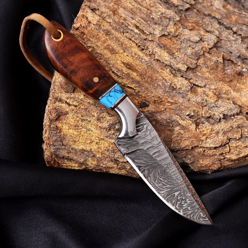 May include: A handcrafted knife with a patterned steel blade and a wooden handle. The handle features a turquoise inlay and a leather lanyard. The knife is displayed on a piece of textured wood, showcasing its craftsmanship and design.