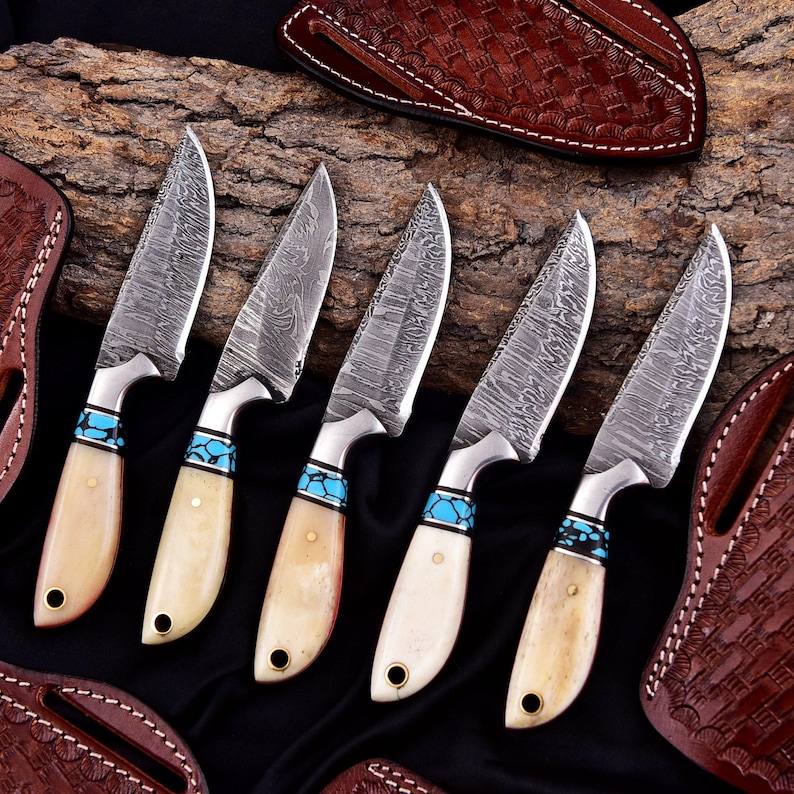 May include: A collection of five Damascus steel knives with bone handles and turquoise accents. The blades have a unique patterned design. Leather sheaths are visible in the background, suggesting a hunting or outdoor theme.