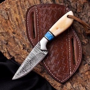 May include: A Damascus steel knife with a bone handle, turquoise and white accents, and a leather sheath. The knife has a curved blade and a lanyard hole. The sheath is brown and embossed with a geometric pattern.
