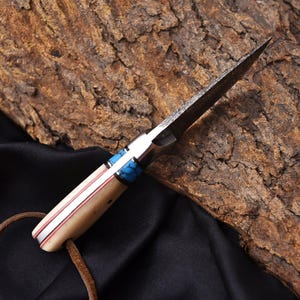 May include: A handcrafted knife with a textured blade and a handle featuring cream-colored bone, red accents, and turquoise inlays. A leather cord is attached to the base of the handle. The knife is set against a dark background.