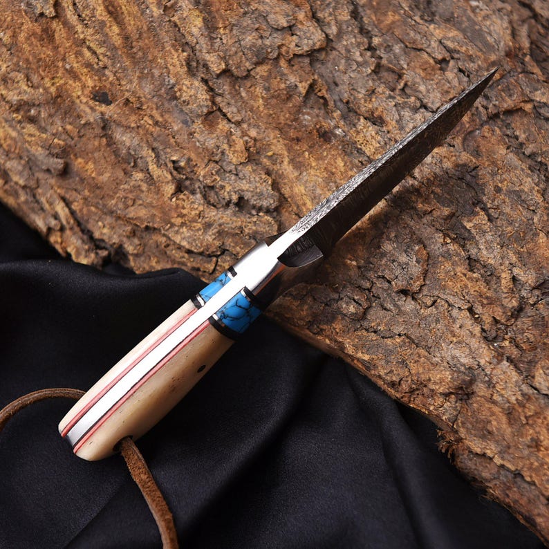 May include: A handcrafted knife with a textured blade and a handle featuring cream-colored bone, red accents, and turquoise inlays. A leather cord is attached to the base of the handle. The knife is set against a dark background.