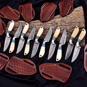 May include: An assortment of handcrafted knives with Damascus steel blades, bone handles, and turquoise accents. The knives are displayed with brown leather sheaths, showcasing a rustic and artisanal aesthetic.