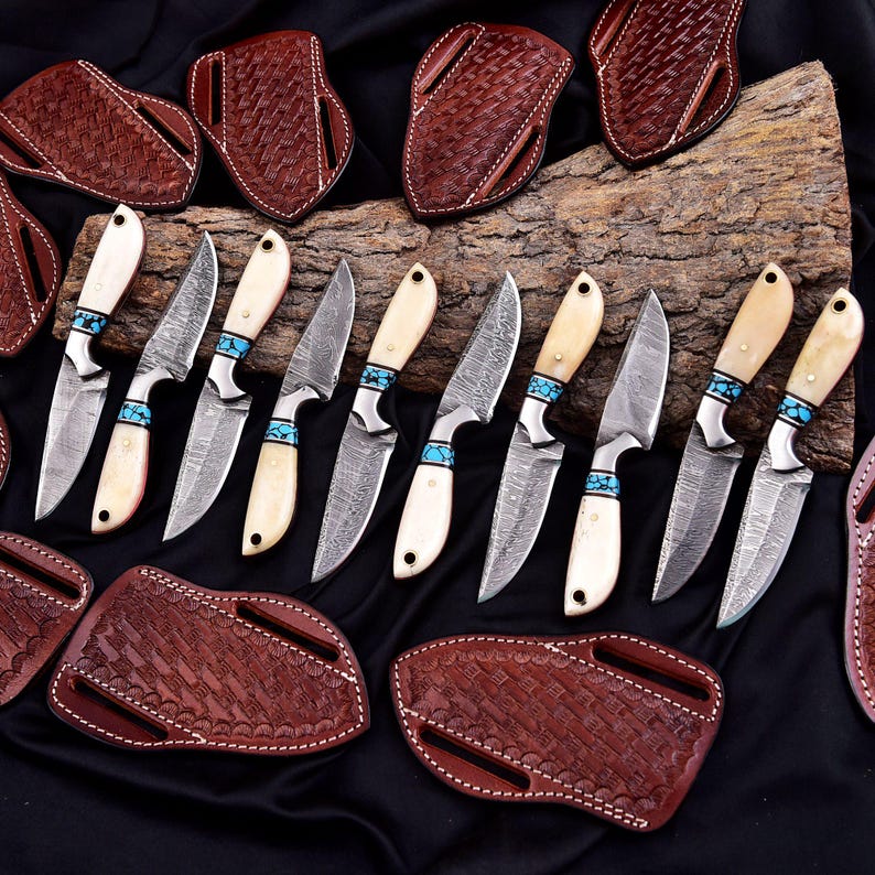 May include: An assortment of handcrafted knives with Damascus steel blades, bone handles, and turquoise accents. The knives are displayed with brown leather sheaths, showcasing a rustic and artisanal aesthetic.
