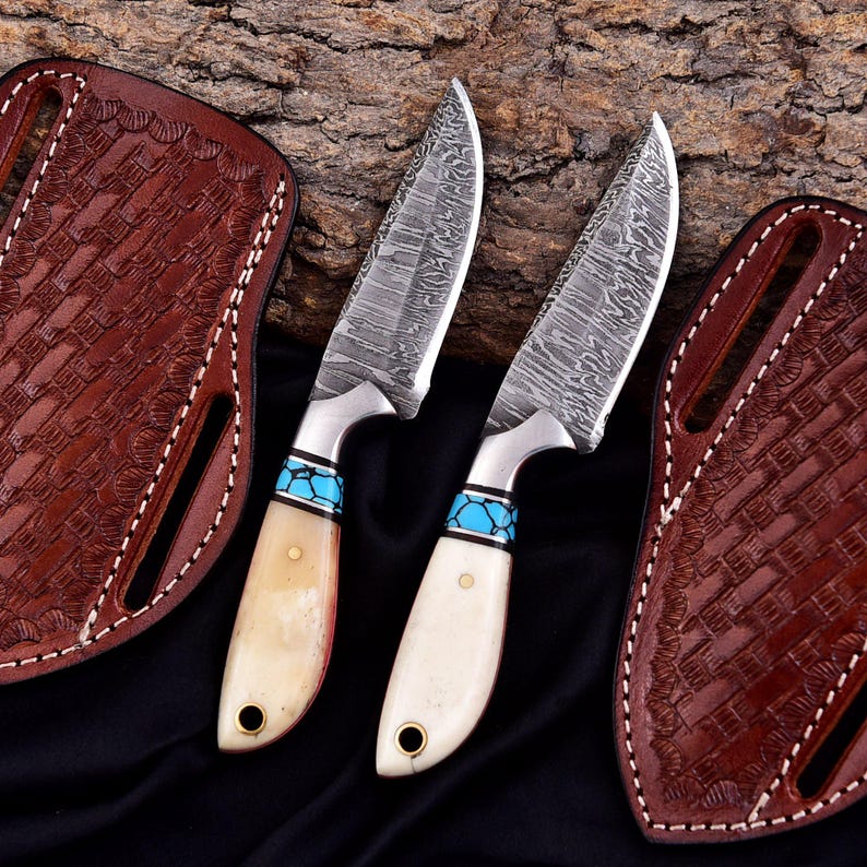 May include: Two Damascus steel knives with bone handles and turquoise accents. The blades have a unique pattern, and the handles are cream-colored. Each knife has a leather sheath with a basket weave design. The sheaths are brown and have white stitching.