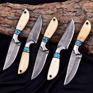 May include: Five handcrafted knives with cream-colored handles and turquoise accents. The blades have a unique patterned design. The knives are arranged on a dark surface with a piece of wood in the background.