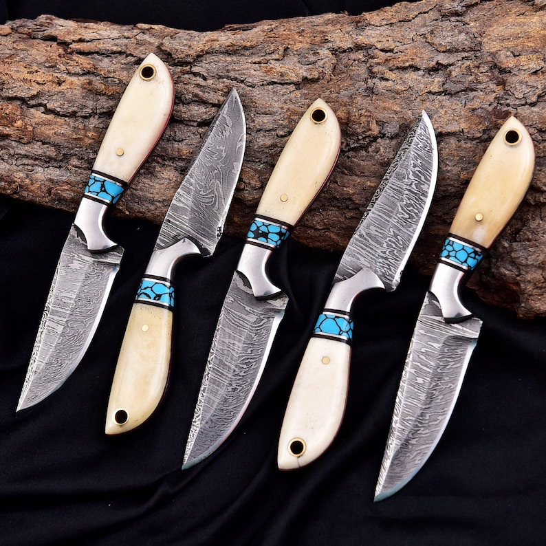 May include: Five handcrafted knives with cream-colored handles and turquoise accents. The blades have a unique patterned design. The knives are arranged on a dark surface with a piece of wood in the background.