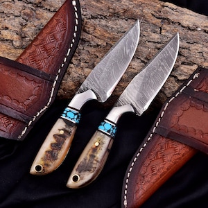 May include: Two Damascus steel knives with bone handles and turquoise accents. The blades have a unique pattern. The handles are attached to the blades with silver bolsters. Brown leather sheaths with decorative stitching are also visible.