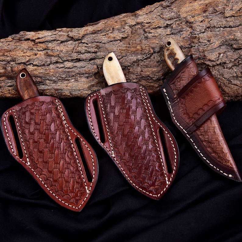 May include: Three handcrafted leather sheaths with knives. The sheaths are a rich brown color with intricate woven patterns and white stitching. The knives have wooden or bone handles. The background includes a piece of wood and a black fabric.