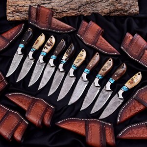 May include: An array of handcrafted knives with patterned blades and various handle designs, including wood and bone, accented with turquoise. Each knife is paired with a brown leather sheath. The knives are arranged on a black surface.