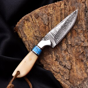 May include: A handcrafted knife with a patterned steel blade and a bone handle. The handle features a blue and silver accent. A leather cord is attached to the base of the handle. The knife is displayed on a piece of wood.