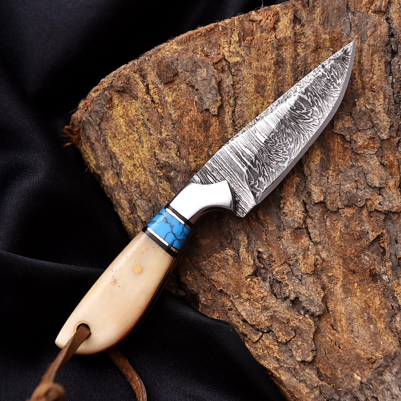May include: A handcrafted knife with a patterned steel blade and a bone handle. The handle features a blue and silver accent. A leather cord is attached to the base of the handle. The knife is displayed on a piece of wood.