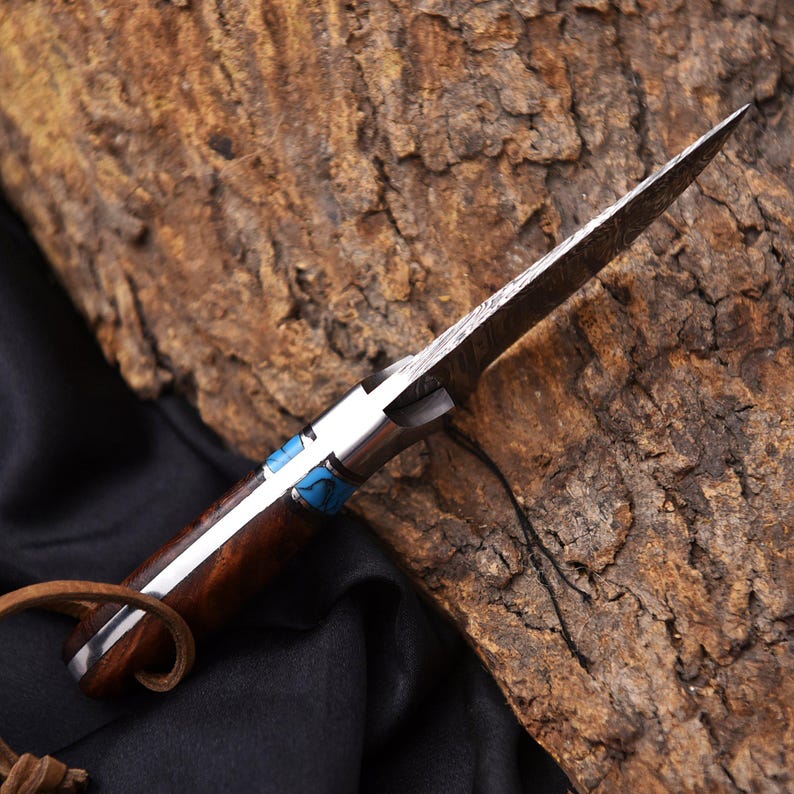 May include: A handcrafted knife with a patterned steel blade and a wooden handle. The handle features a silver bolster and blue accents. A leather loop is attached to the end of the handle.