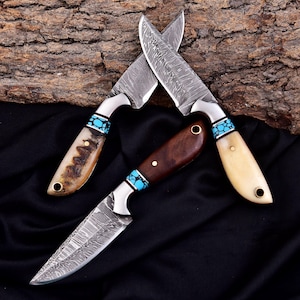 May include: Three handcrafted knives with patterned steel blades. The handles feature wood, bone, and turquoise accents. The blades are crossed, showcasing the intricate designs and materials used in their construction.