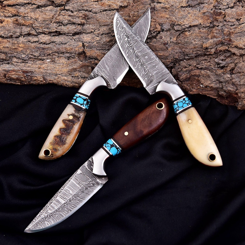 May include: Three handcrafted knives with patterned steel blades. The handles feature wood, bone, and turquoise accents. The blades are crossed, showcasing the intricate designs and materials used in their construction.