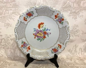 Peut inclure: Une assiette décorative en porcelaine blanche avec un bord festonné et une bordure dorée. L'assiette présente un motif floral au centre et sur le bord. Les fleurs sont dans les tons de rouge, violet, jaune et bleu. L'assiette est présentée sur un support noir.