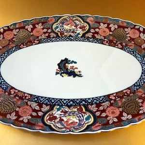 May include: An oval-shaped ceramic serving platter with a white center and a decorative border. The border features a maroon background with floral and bird motifs in shades of pink, blue, and gold. The platter measures approximately 16 inches in length.