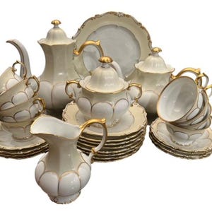 May include: A complete antique tea set in cream with gold trim. The set includes teacups, saucers, a teapot, a sugar bowl, a creamer, and serving plates. The edges of the plates and cups have a scalloped design.