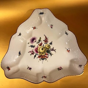 Op de afbeelding: Een witte, driehoekige serveerschaal met een geschulpte rand en gouden rand. De schaal is versierd met een centraal bloemstuk in tinten geel, roze, paars en groen, met kleinere bloemaccenten rond de randen.