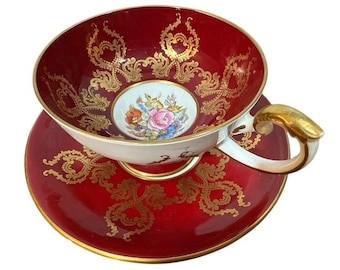 May include: A red teacup and saucer set with gold filigree detailing. The teacup interior and saucer are red, with gold scrollwork. The teacup features a floral design in the center. The handle and rim are gold.