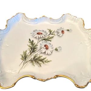 May include: An off-white porcelain serving tray with a gold trim and handles. The tray features a floral design of white daisies with brown centers and green leaves. The edges have a scalloped design.